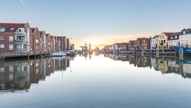 Ein Kanal, gesäumt von Gebäuden, mit wolkenlosem Himmel und untergehender Sonne in der Ferne.