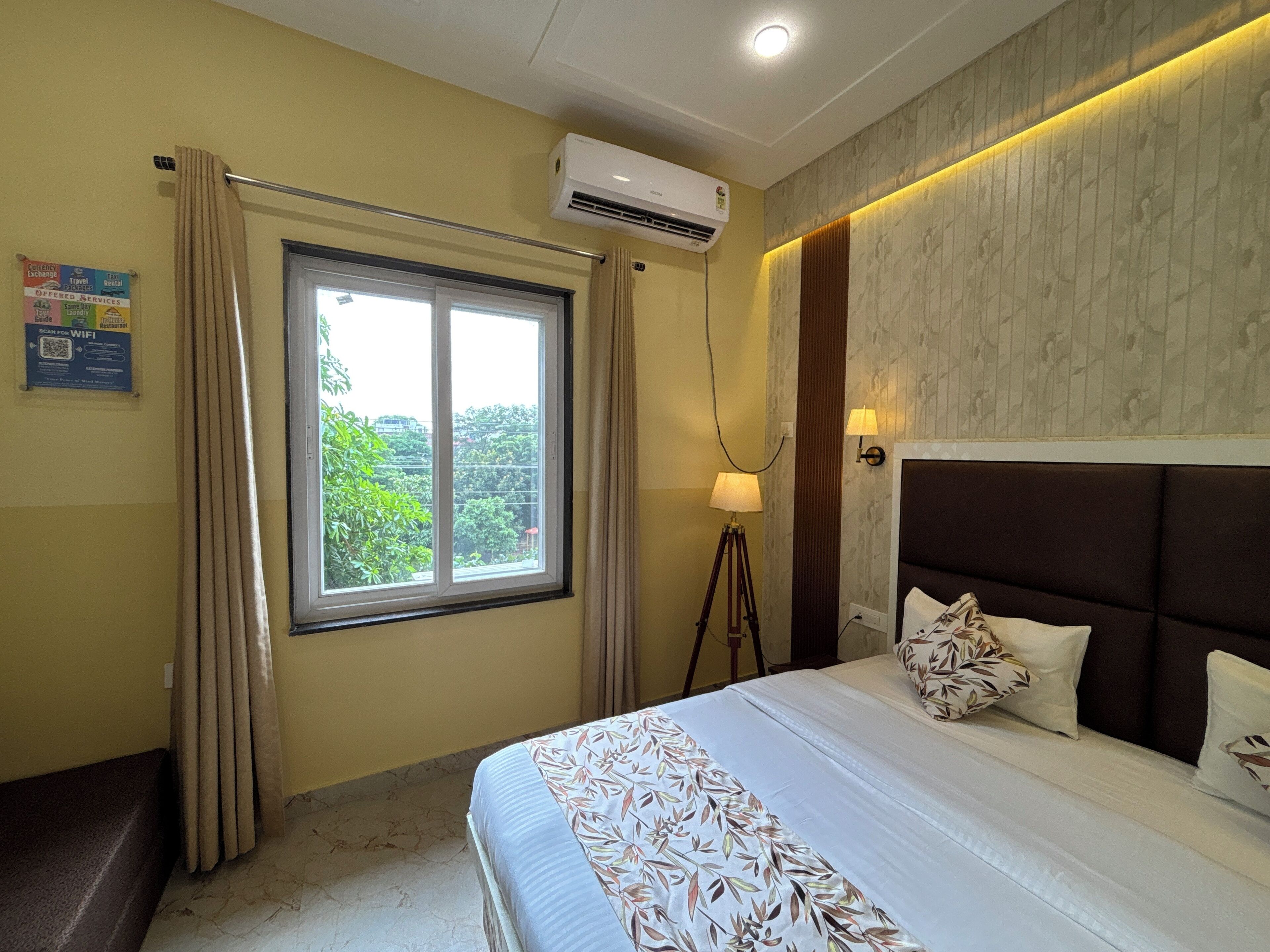 Een hotelkamer met een bed, een raam met gordijnen, een airconditioner, een lamp en een wandlamp.