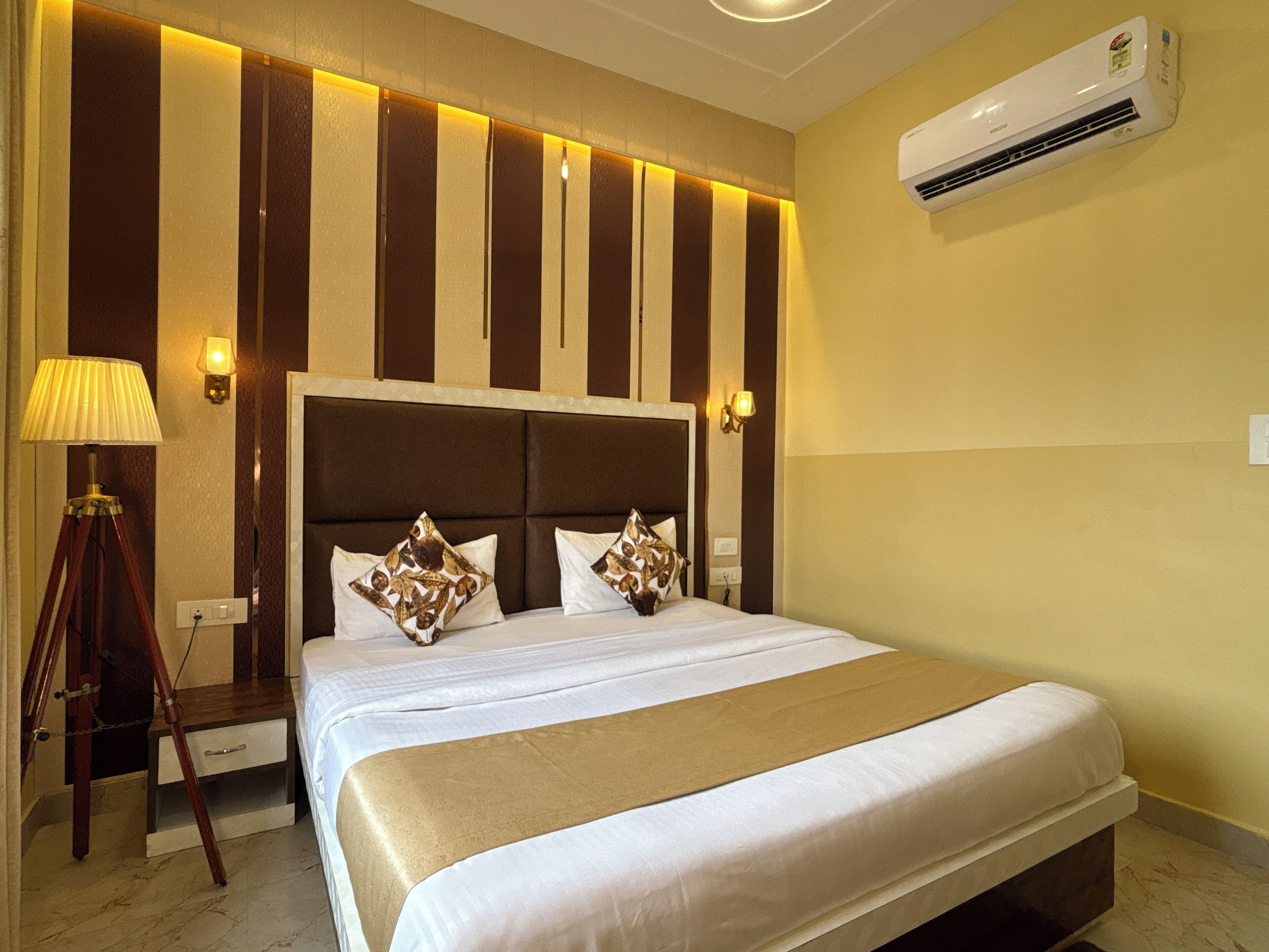 A hotel room with a large bed, a wooden nightstand, a lamp, and an air conditioning unit.