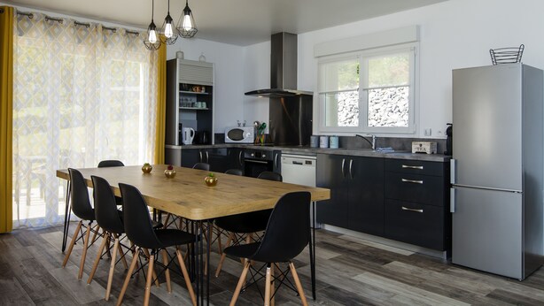 A modern kitchen with a wooden dining table, black chairs, stainless steel appliances, and a window with a view of trees.