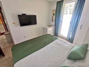 Un dormitorio con cama, televisor en la pared, una mesita de noche pequeña y una ventana con cortinas.