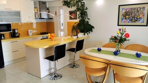 A modern kitchen with a yellow countertop, two black bar stools, a white dining table with a green placemat, and a potted plant. - Mario` place - penthouse with sea view terrace (Dubrovnik)