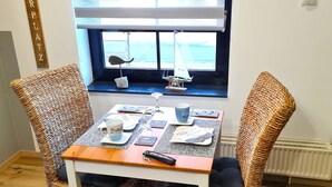 Een eetgedeelte met een tafel gedekt voor twee personen, rieten stoelen, een raam met zonwering en een klein model zeilbootje op de vensterbank.