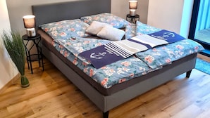 Een moderne slaapkamer met een grijs gestoffeerd bed, bloemenbeddengoed en uitzicht op het landschap door een raam.