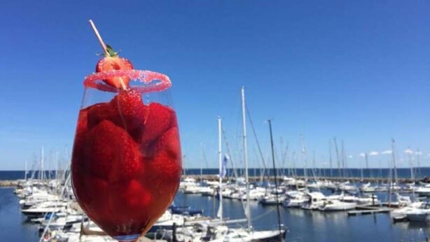 Een glas rode drank met een aardbeien garnering en rietje, voor een achtergrond van een jachthaven met boten.