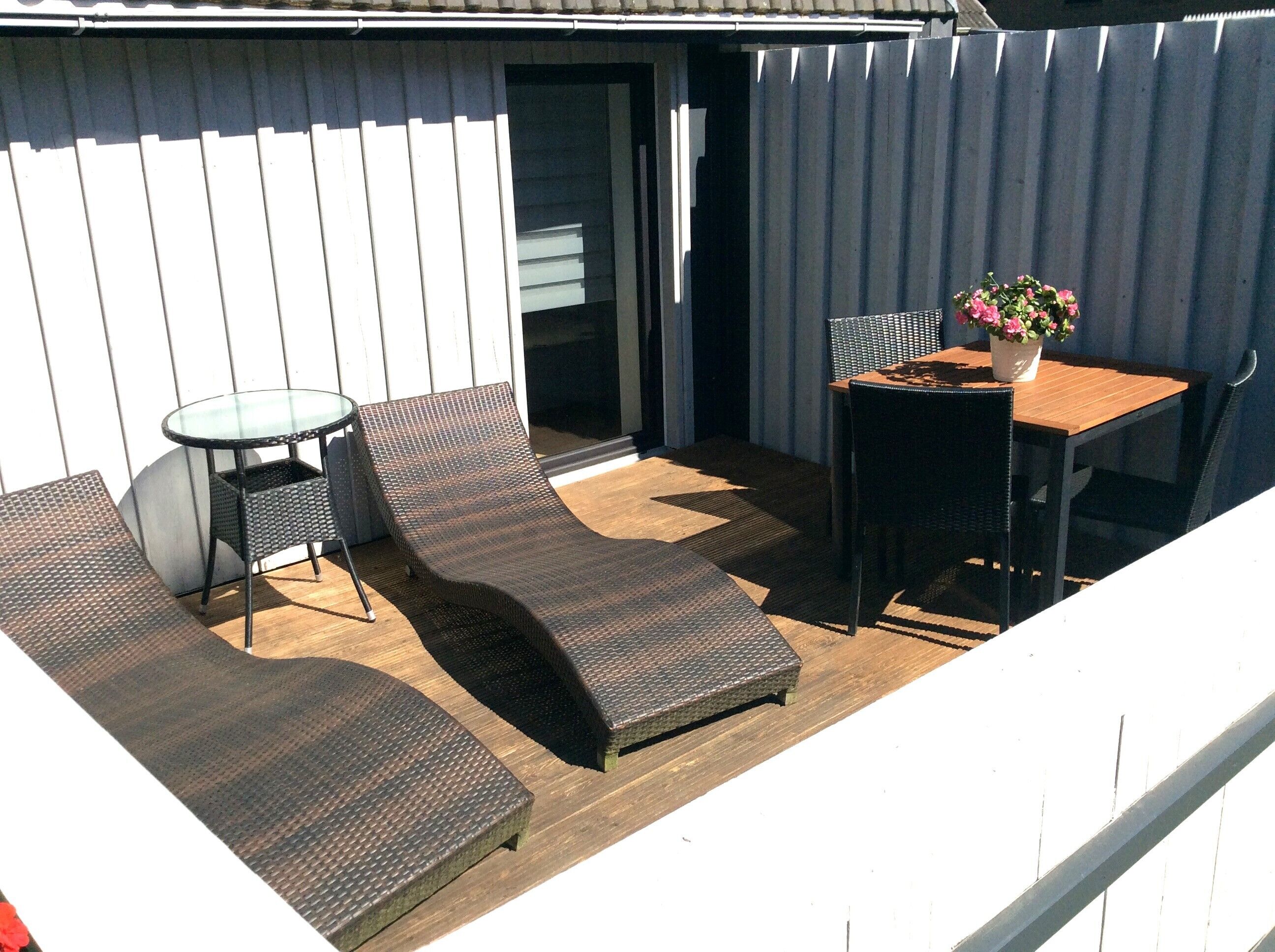 A patio with a glass table, two cushioned chairs, and a wooden table with a potted plant.