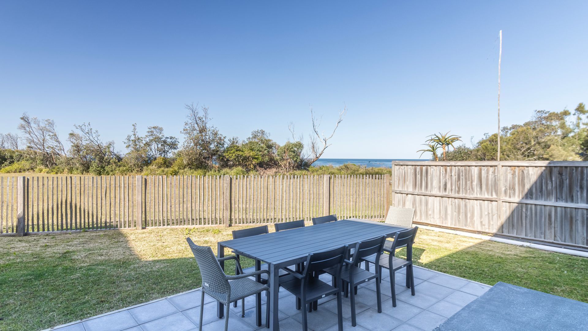 An outdoor patio with a table and chairs, a wooden fence, and a view of the ocean.