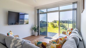 A modern living room with a large sliding glass door leading to a garden, a flat-screen TV mounted on the wall, and a cozy seating area with patterned cushions. - Beachside Cottage - Cudmirrah (Cudmirrah)