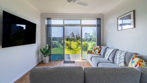 A modern living room with a grey sofa, a flat-screen TV, and a large sliding glass door leading to a garden. - Beachside Cottage - Cudmirrah (Cudmirrah)