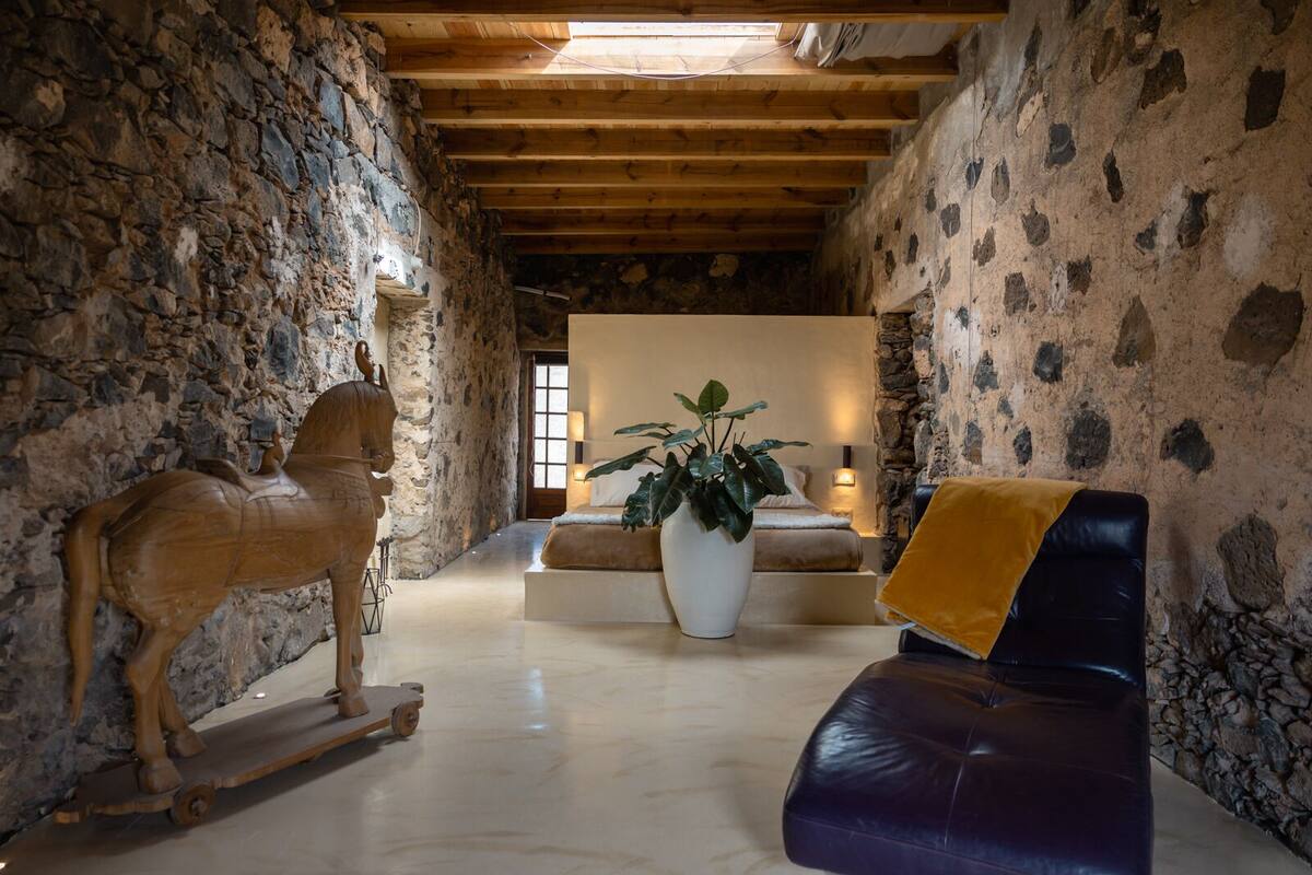 Un interior rústico con paredes de piedra, una escultura de madera de un caballo, una planta grande en maceta y un sofá de cuero.