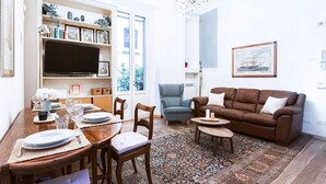 A living room with a brown sofa, a chandelier, a television, and a dining area with a table set for four. - Easylife - Prestigious apartment in Risorgimento (Milano)