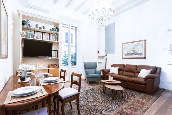 A living room with a brown sofa, a chandelier, a television, and a dining area with a table set for four. - Easylife - Prestigious apartment in Risorgimento (Milano)