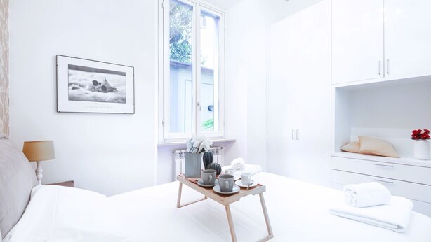 A bright, white room with a bed, a bedside table with a lamp, a window, a framed picture on the wall, and a small wooden table with cups and a teapot. - Easylife - Prestigious apartment in Risorgimento (Milano)