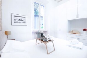 A bright, white room with a bed, a bedside table with a lamp, a window, a framed picture on the wall, and a small wooden table with cups and a teapot. - Easylife - Prestigious apartment in Risorgimento (Milano)