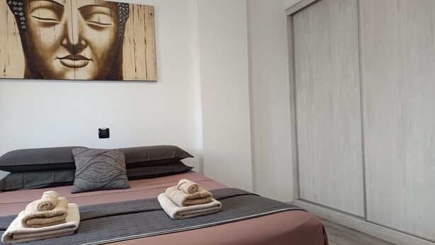 Ein modernes Schlafzimmer mit einem großen Bett, dunkler Bettwäsche und einem an der Wand befestigten Buddha-Gemälde.