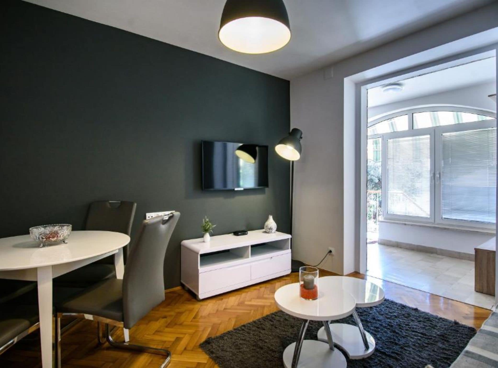 Una sala de estar moderna con una mesa redonda de comedor, un mueble blanco, un televisor montado en la pared y un amplio ventanal con persianas.