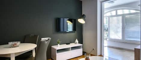 Ein modernes Wohnzimmer mit einem runden Esstisch, einem weißen Schrank, einem an der Wand montierten Fernseher und einem großen Fenster mit Jalousien.