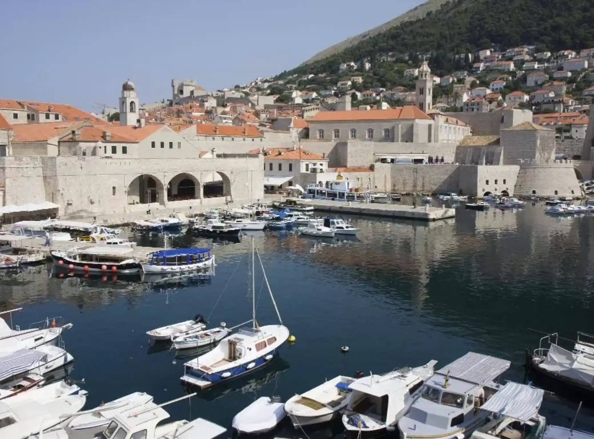 A marina with boats docked, a historic building with arches, and a hillside town in the background.