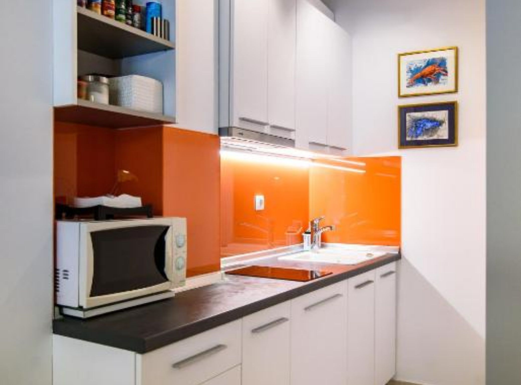 A modern kitchen with white cabinets, an orange accent wall, a microwave, and a sink.