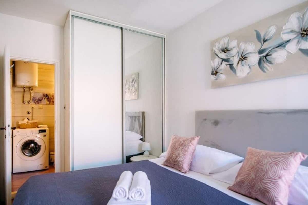 Ein Hotelzimmer mit Bett, Waschmaschine, Spiegel und einem floralen Bild an der Wand.