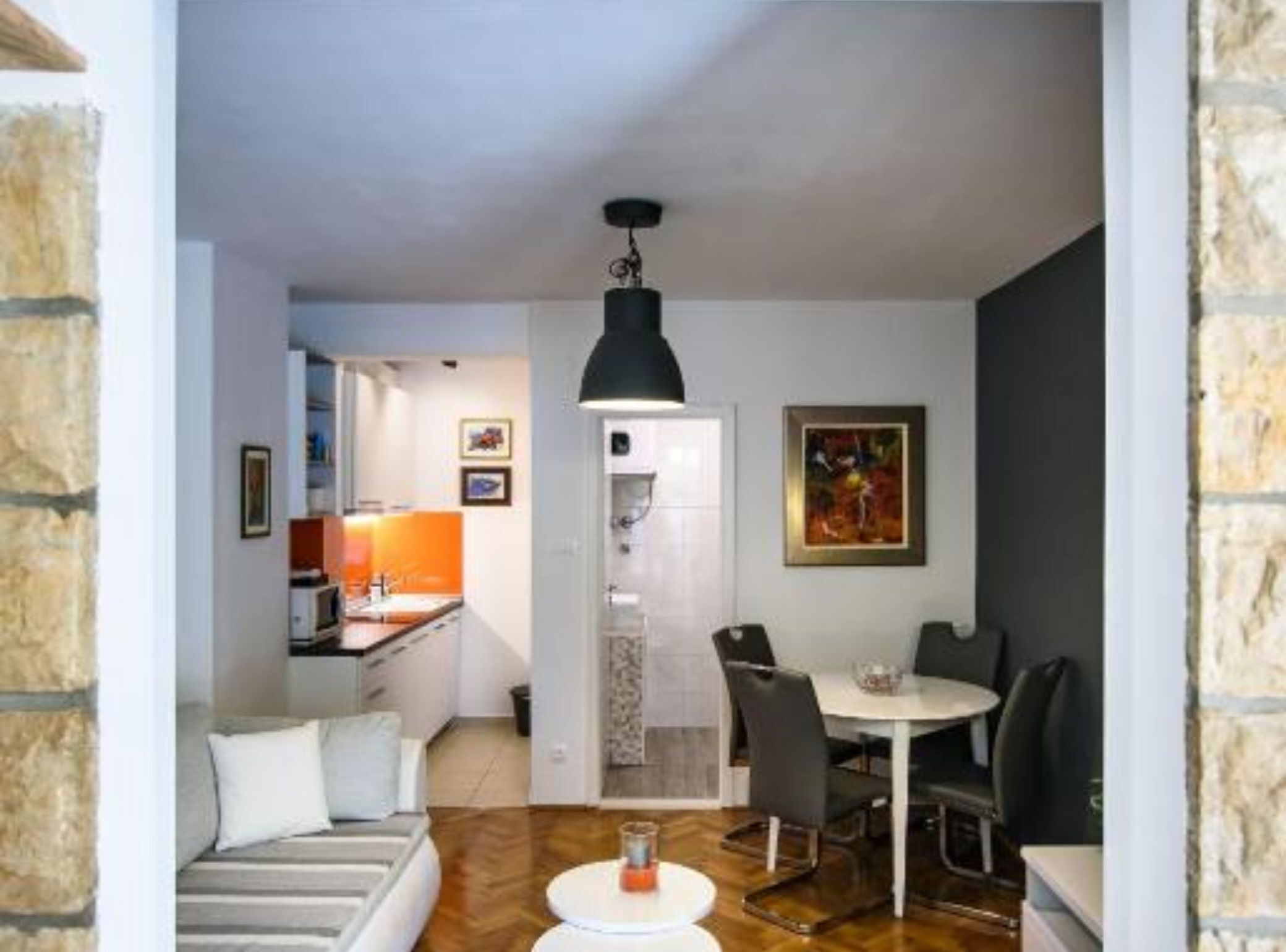 A modern living room with a sofa, dining table, and chairs. There is a kitchen area with a sink and a refrigerator. A painting is on the wall, and a ceiling light fixture is visible.