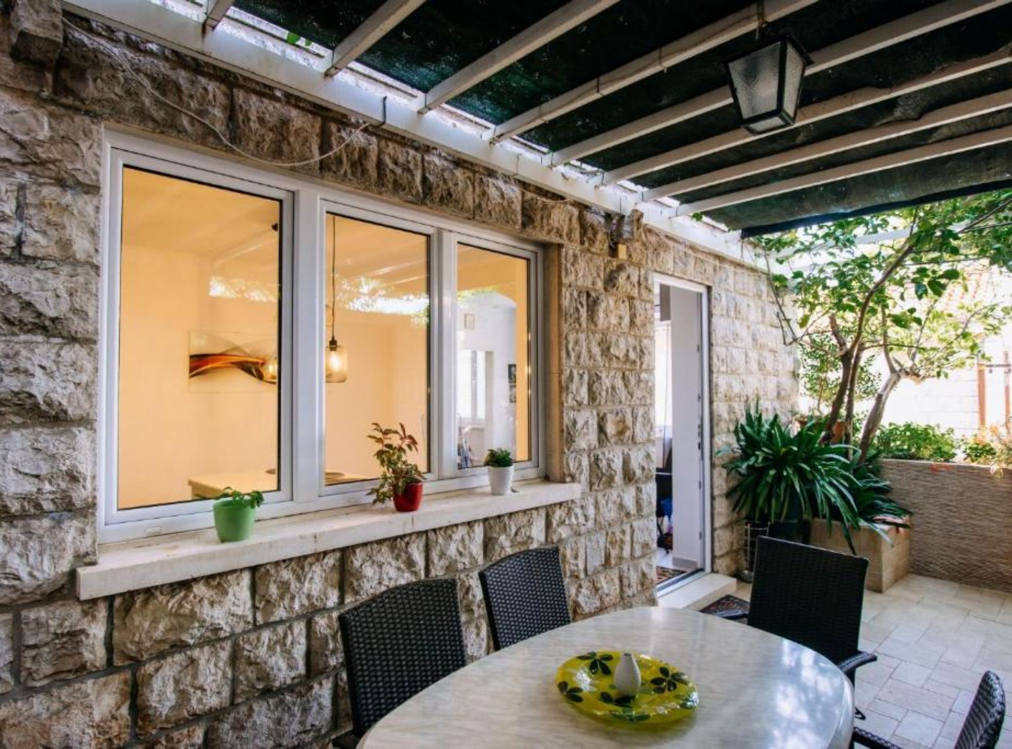 A stone exterior wall with large windows, a covered patio area with a round table and chairs, and potted plants.