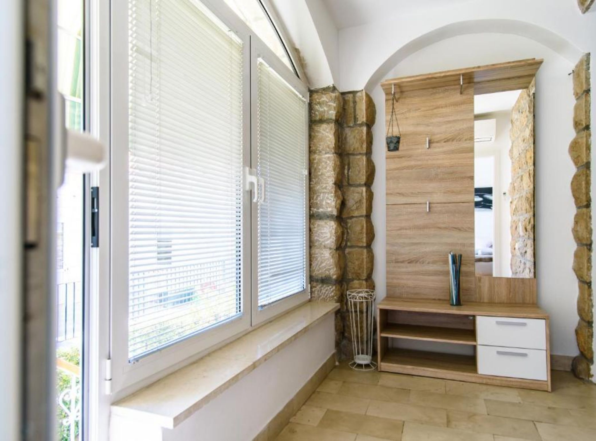 A room with a large window, stone wall, wooden cabinet, and a small trash can.