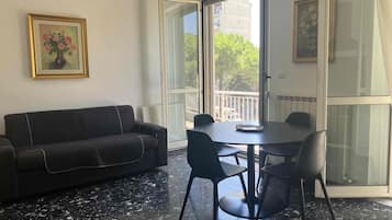 Ein Zimmer mit einem dunklen Sofa, einem runden Tisch mit Stühlen, einem großen Fenster und einem gerahmten Bild an der Wand.