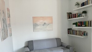 A grey sofa with a textured blanket, a wall-mounted painting, and a bookshelf with books and plants.