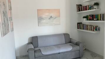 Ein graues Sofa mit einer gemusterten Decke, ein Wandbild und ein Bücherregal mit Büchern und Pflanzen.