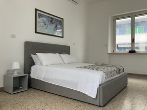 Ein Schlafzimmer mit grauem Bett, weißer Bettwäsche, einem kleinen Nachttisch mit Lampe, einem gerahmten Kunstwerk an der Wand und einem Fenster mit Blick auf Gebäude.