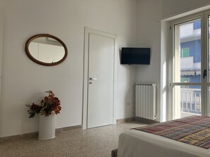 A bedroom with a bed, a white door, a round mirror, a TV, a radiator, and a window with a view of buildings.