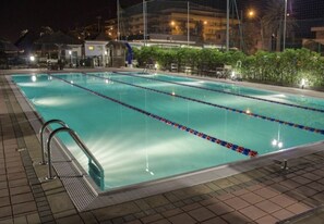 Ein Schwimmbecken mit mehreren Bahnen, umgeben von einer gefliesten Terrasse und einem Zaun.