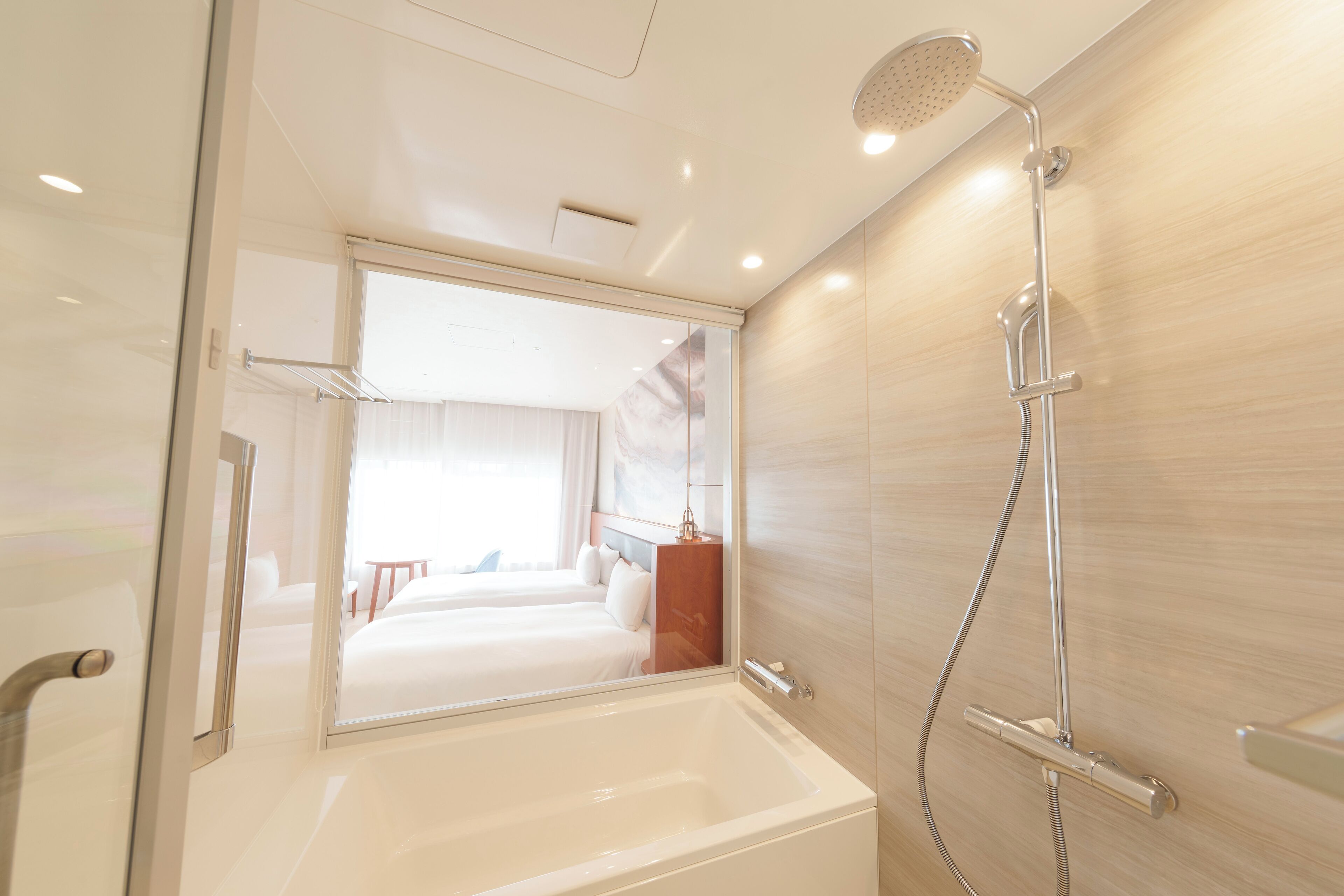Un baño moderno con una bañera amplia, un cabezal de ducha y un espejo que refleja un dormitorio con una cama y una mesita de noche.