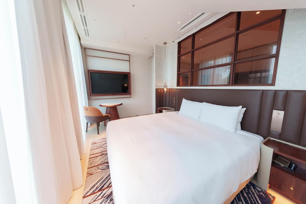 Ein Hotelzimmer mit einem großen Bett, einem an der Wand montierten Fernseher, einem kleinen Tisch und einem Fenster mit Vorhängen.