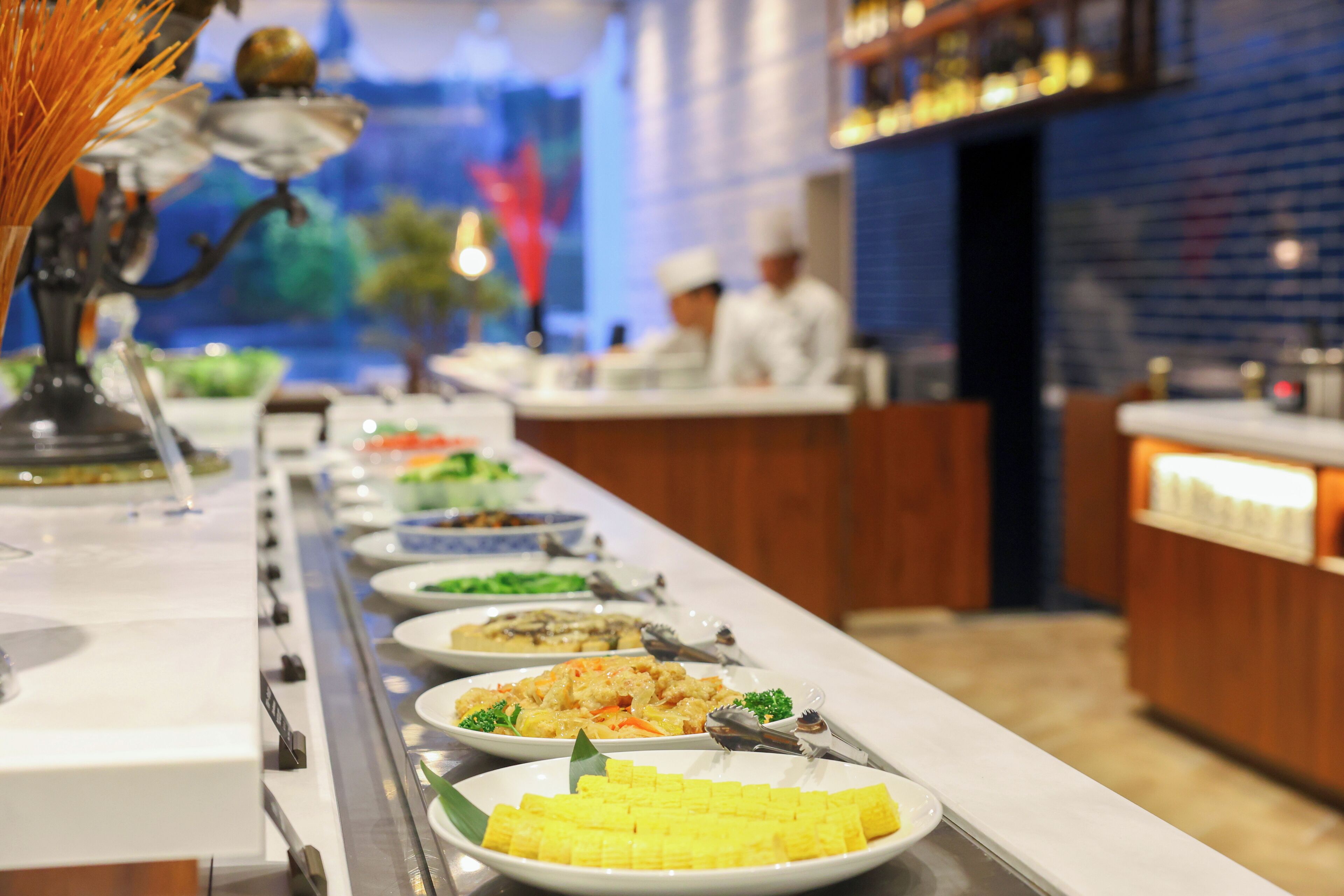 Een buffet met diverse gerechten, een decoratieve vaas en een kok op de achtergrond.