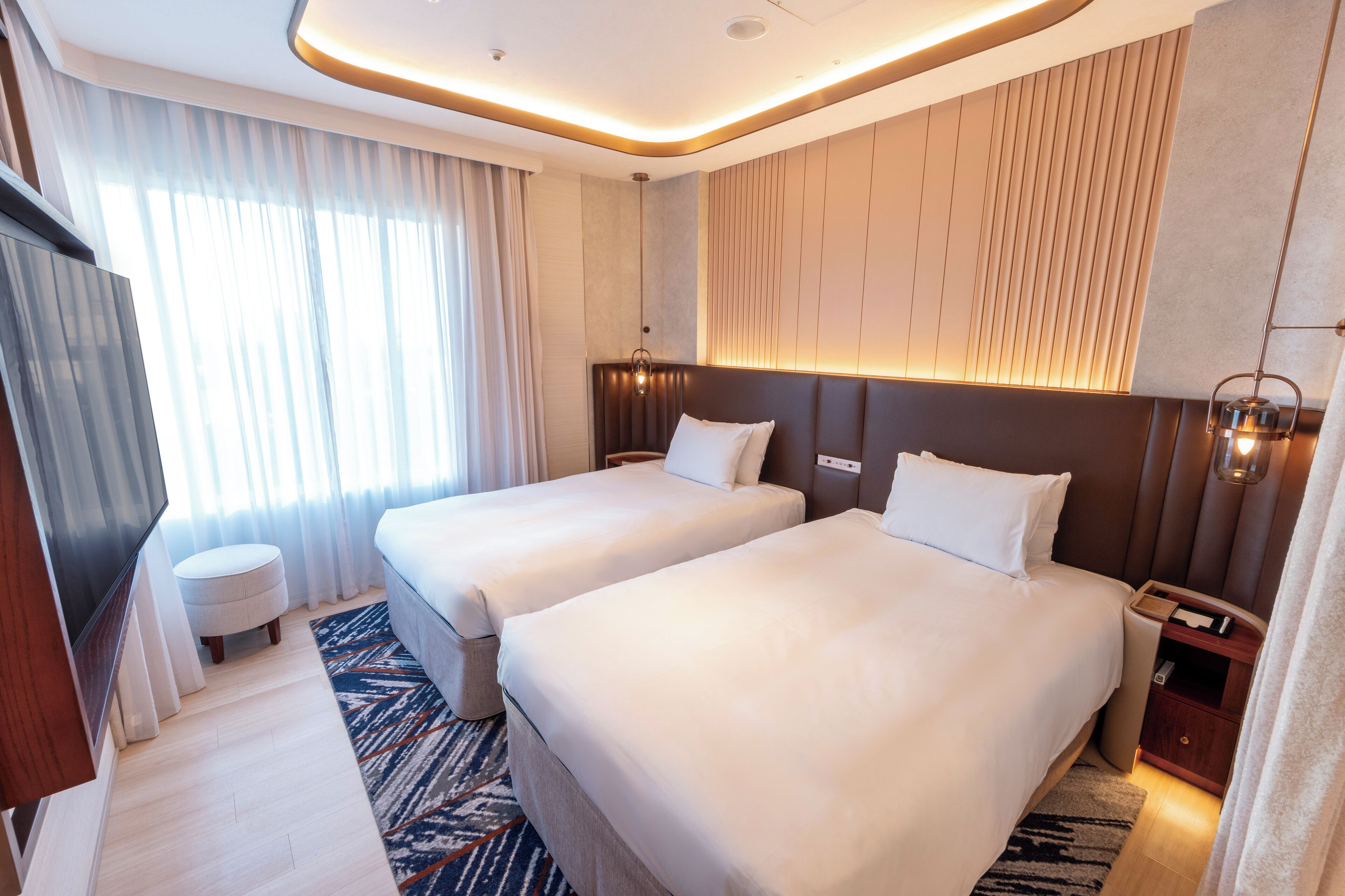 Quarto de hotel com duas camas, televisão, mesa de cabeceira e uma janela grande com cortinas.
