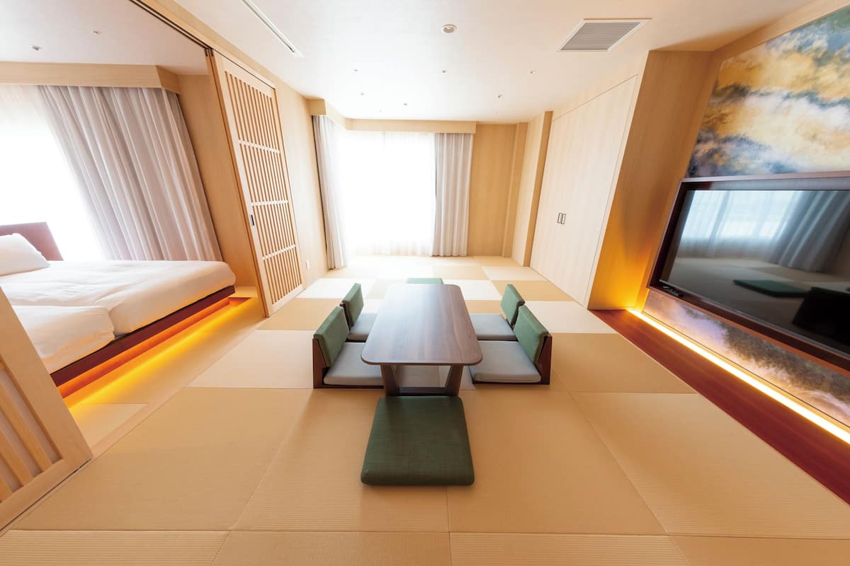 Ein modernes Hotelzimmer mit Bett, Esstisch mit Bänken, Fernseher und großen Fenstern mit Vorhängen.
