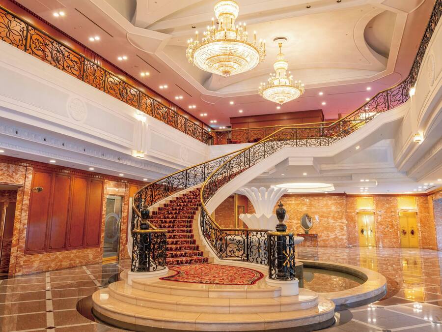 Un lobby imponente con piso de mármol, una gran escalera con barandillas ornamentadas y candelabros.