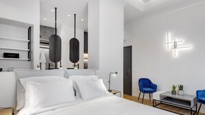 A modern hotel room with a large bed, a desk, two blue chairs, a small table with a plant, and a wall-mounted light fixture. - Times Elegant Rooms (Syros)