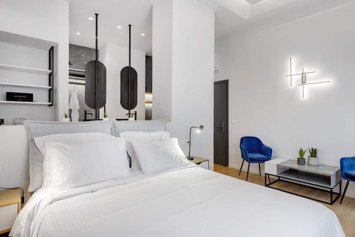 Una habitación de hotel moderna con una cama grande, un escritorio, dos sillas azules, una mesita con una planta y una lámpara de pared.