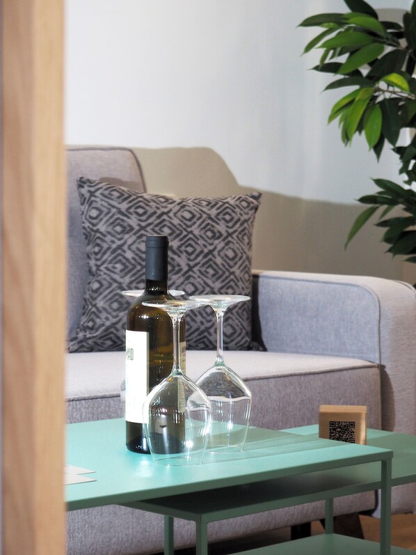 Mesa com uma garrafa de vinho e dois copos de vinho, almofada estampada em um sofá e uma planta ao fundo.
