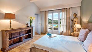 A bedroom with a bed, a wooden side table with a lamp, a statue, and a view of a swimming pool through a glass door.