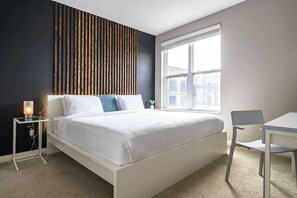 Un dormitorio moderno con cama, mesita de noche, lámpara y ventana con vista a edificios.