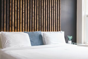 A bed with white linens and a blue textured pillow, a small potted plant on a bedside table, and a wooden slat wall behind the bed.