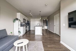 A modern apartment with a grey sofa, a small white table, and a kitchen area with white cabinets and a central island.