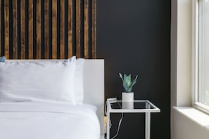 Una camera d'hotel moderna con una parete in listelli di legno, un letto bianco, un comodino bianco e piccolo con una pianta in vaso, e una finestra con vista.