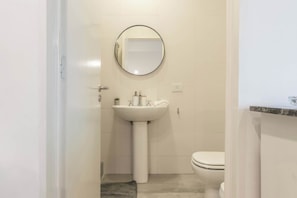 Une petite salle de bain avec un lavabo blanc, un miroir rond et des toilettes.