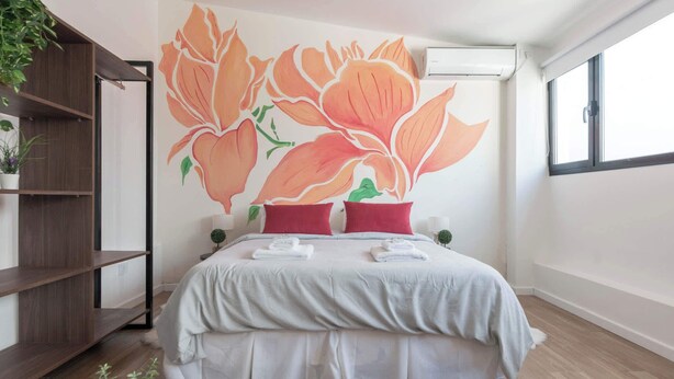 A bedroom with a bed, pillows, a shelf, a potted plant, and a wall mural.
