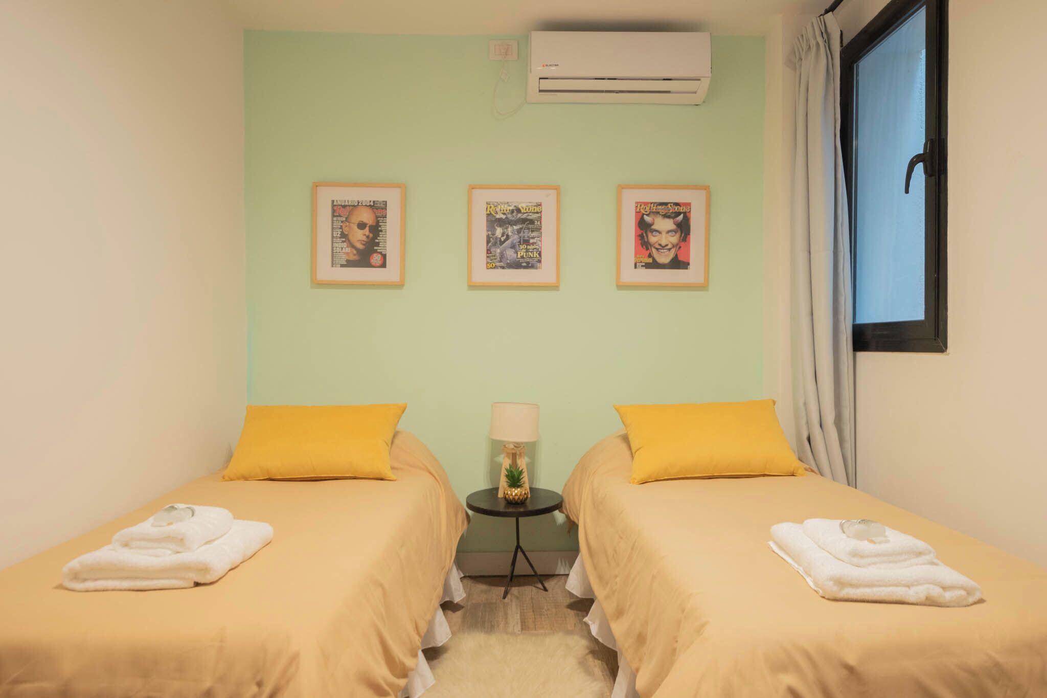 A room with two beds, each with a yellow pillow and a white towel, a small table with a lamp, and three framed pictures on the wall.